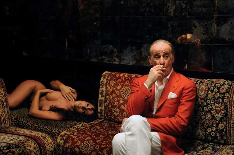 Toni Servillo como Jep Gambardella, de blazer vermelho, fuma num sofá ornamentado enquanto uma mulher descansa ao lado