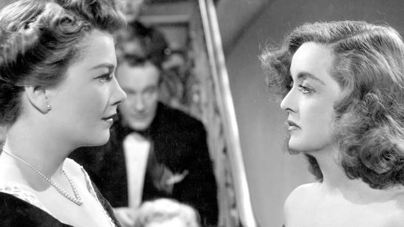 Anne Baxter e Bette Davis frente a frente em cena em preto e branco, com um homem de smoking ao fundo