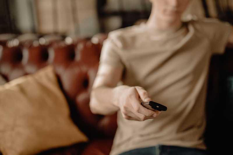 Homem no sofá de couro apontando controle remoto preto para a câmera, com fundo desfocado