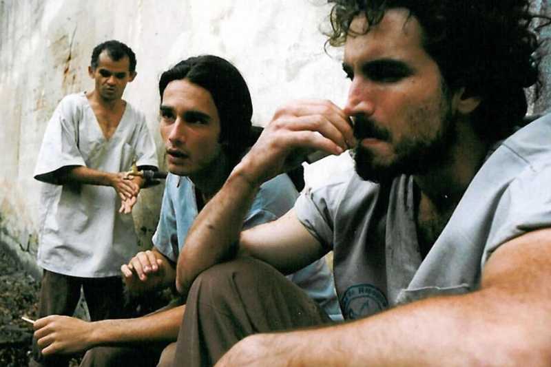 Três rapazes de roupas claras junto a um muro descascado; o da frente, de barba, leva a mão ao rosto