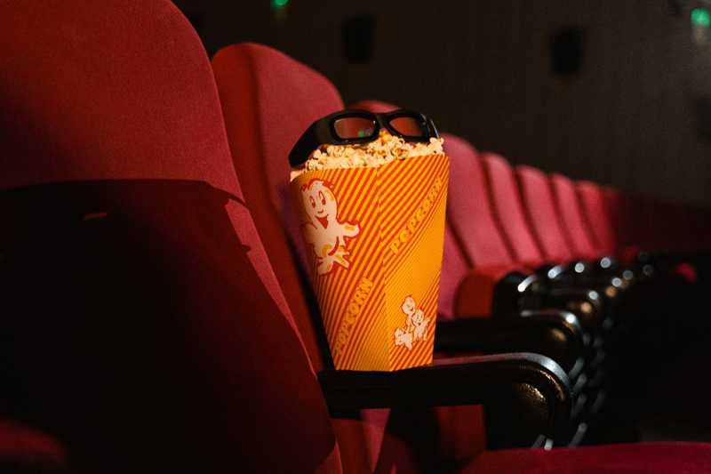 Balde de pipoca com óculos 3D sobre o apoio de braço de poltrona vermelha em sala de cinema