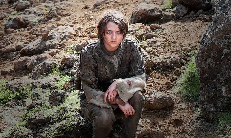 Maisie Williams como Arya Stark, sentada entre pedras numa encosta