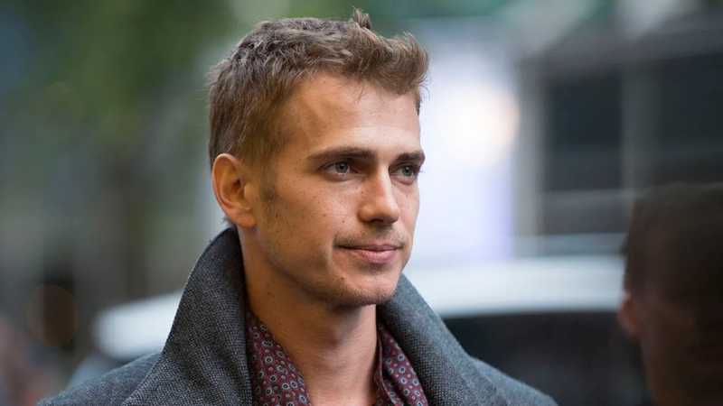 Hayden Christensen de sobretudo cinza e camisa estampada, em close com leve sorriso e fundo desfocado