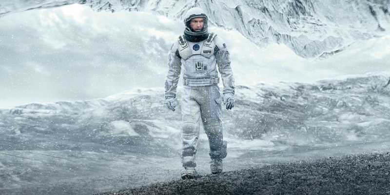 Cooper de traje espacial branco em planeta gelado, com formações de gelo ao fundo e neve no ar