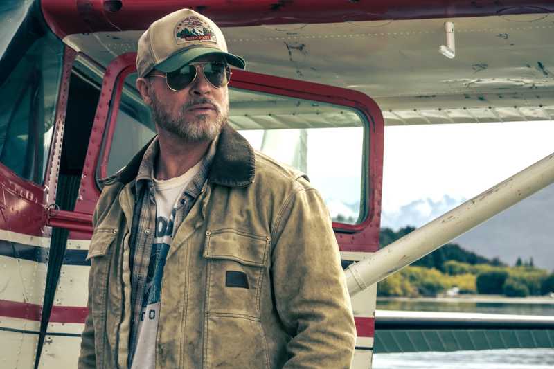 Homem de boné, óculos de aviador e jaqueta de lona ao lado de um hidroavião, com lago e montanhas ao fundo