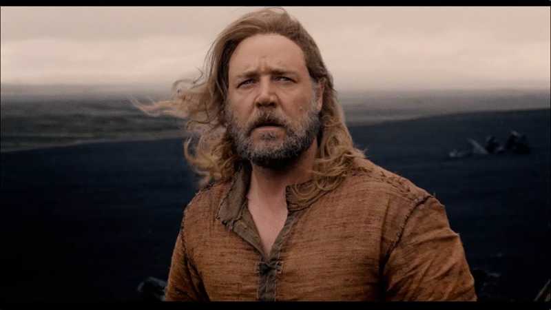 Russell Crowe como Noé, de barba grisalha e túnica rústica, numa paisagem desolada sob céu nublado