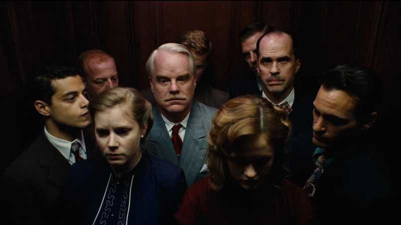 Philip Seymour Hoffman e Amy Adams, cercados por outros personagens, dentro de um elevador