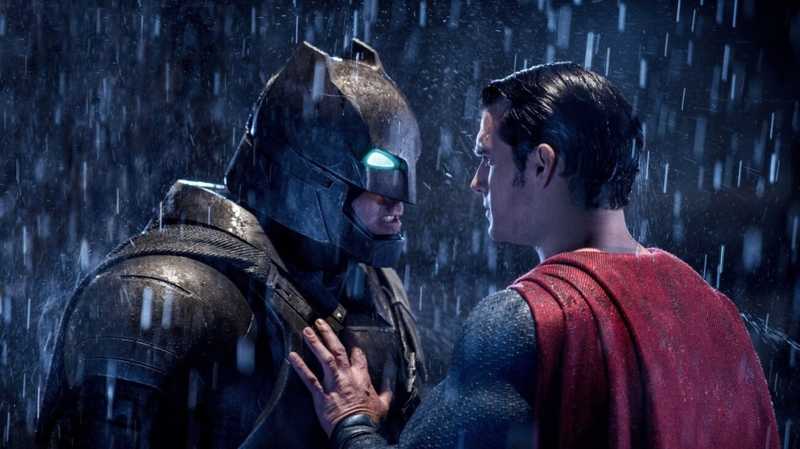 Batman de armadura metálica encara Superman de perfil sob chuva intensa, mão no peito do herói