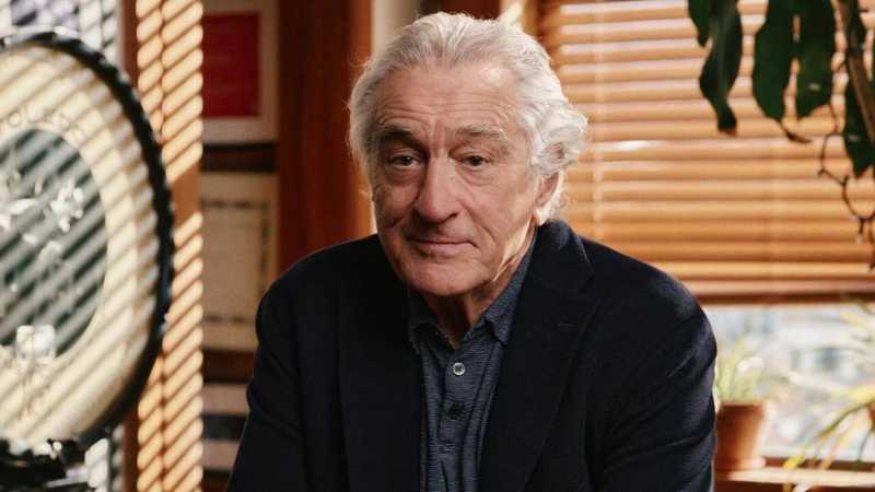 Robert De Niro de blazer azul-marinho e polo listrado, olhando para a câmera com leve sorriso em ambiente iluminado