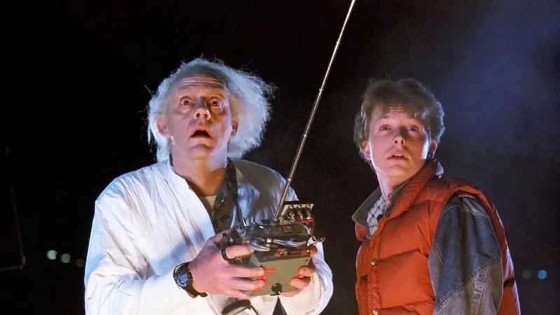 Doc Brown de macacão branco com controle remoto e Marty McFly de colete laranja olham para cima, surpresos, à noite