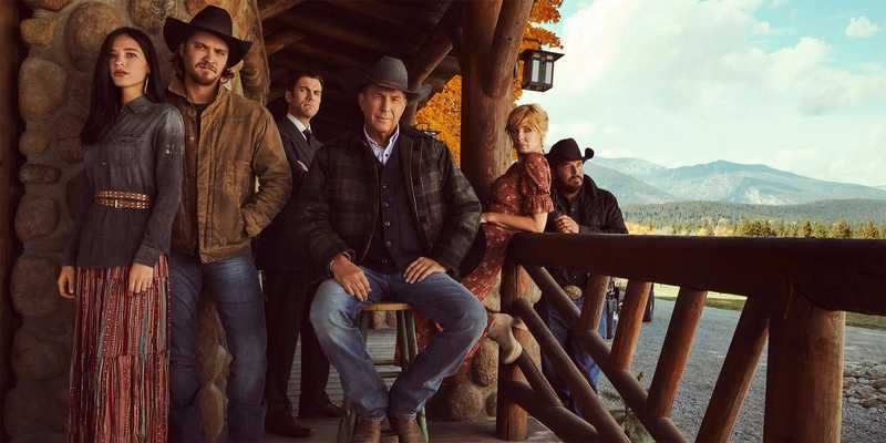 Elenco principal de Yellowstone posando na varanda do rancho Dutton com as montanhas de Montana ao fundo